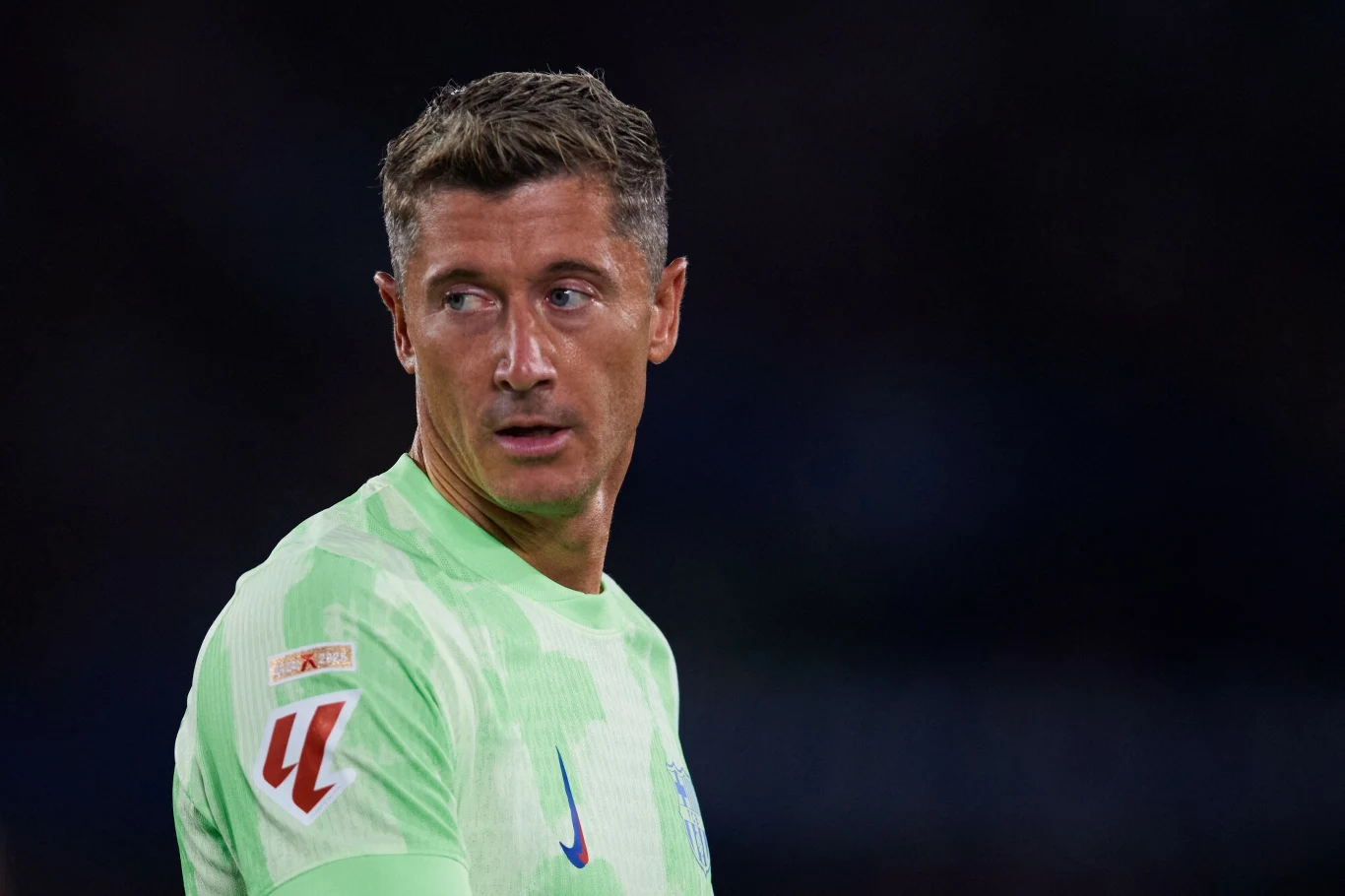 Piłkarz w jasnozielonej koszulce sportowej spogląda przez ramię, tło ciemne i rozmyte, twarz dobrze widoczna z wyrazem skupienia.