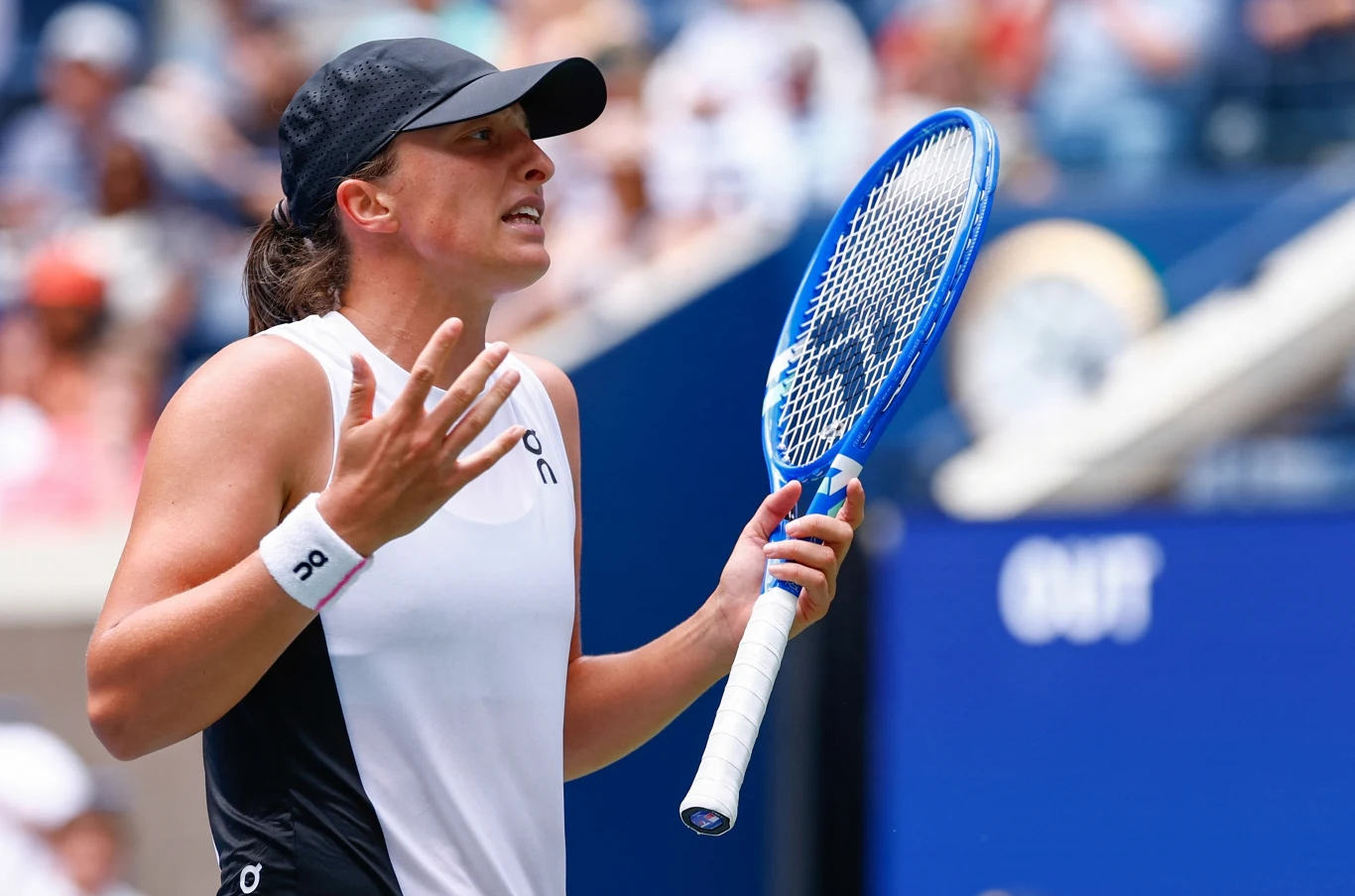 Tenisistka w sportowym stroju i czapce trzymająca rakietę, gestykulująca obiema dłońmi w geście wyrażającym emocje, tło rozmyte z widownią oraz elementami kortu tenisowego.