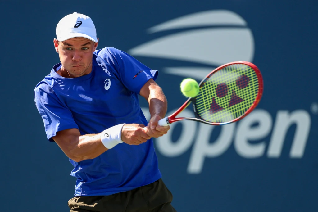 Tenisista w niebieskiej koszulce i białej czapce uderza piłkę tenisową z backhandu na korcie, w tle widoczne logo turnieju US Open.