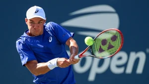 Tenisista w niebieskiej koszulce i białej czapce uderza piłkę tenisową z backhandu na korcie, w tle widoczne logo turnieju US Open.