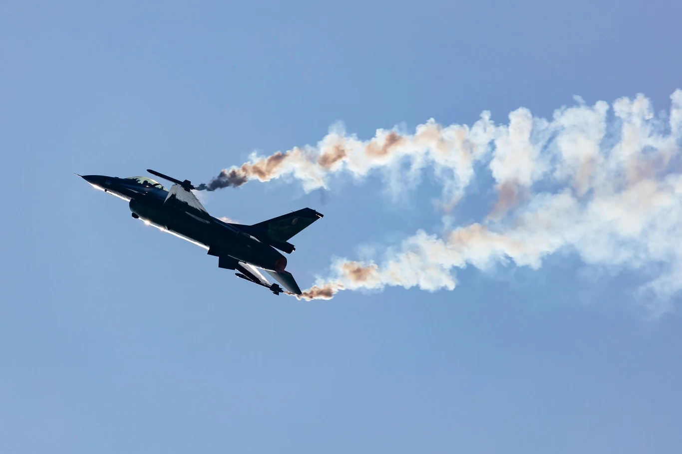 Pokaz F-16 w Radomiu (zdj. arch.) Samolot myśliwski wykonujący akrobację na niebie pozostawiający za sobą smugi dymu na tle błękitnego nieba.