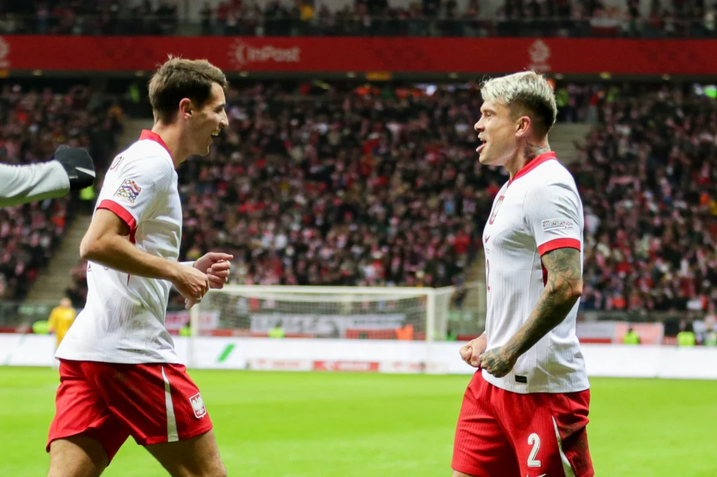 Kamil Piątkowski i Jakub Kiwior Dwóch piłkarzy w biało-czerwonych strojach świętuje sukces podczas meczu na stadionie, w tle rozmyty tłum kibiców na trybunach.