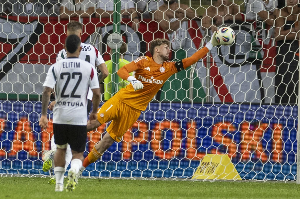 Bramkarz w pomarańczowym stroju wykonuje efektowną paradę, próbując obronić strzał piłki kierującej się w stronę bramki, piłkarze w białych koszulkach obserwują akcję, w tle widoczna siatka bramkowa oraz kibice.