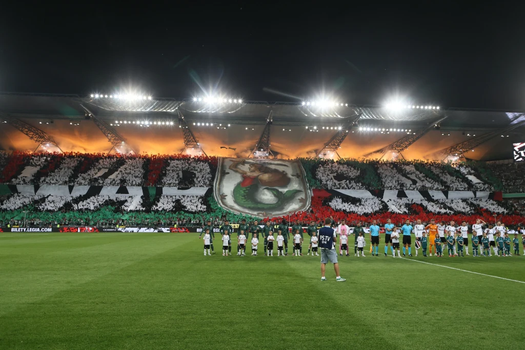 Tłum kibiców na stadionie, tworzący wielką oprawę z napisem 'WARSZAWA' oraz herbem klubu, przed nimi na murawie ustawieni zawodnicy obu drużyn i sędziowie, jasno oświetlone trybuny na tle wieczornego nieba.