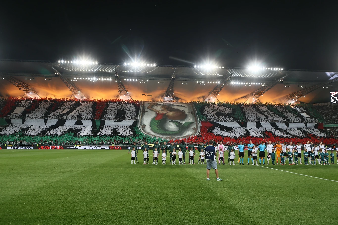Tłum kibiców na stadionie, tworzący wielką oprawę z napisem 'WARSZAWA' oraz herbem klubu, przed nimi na murawie ustawieni zawodnicy obu drużyn i sędziowie, jasno oświetlone trybuny na tle wieczornego nieba.