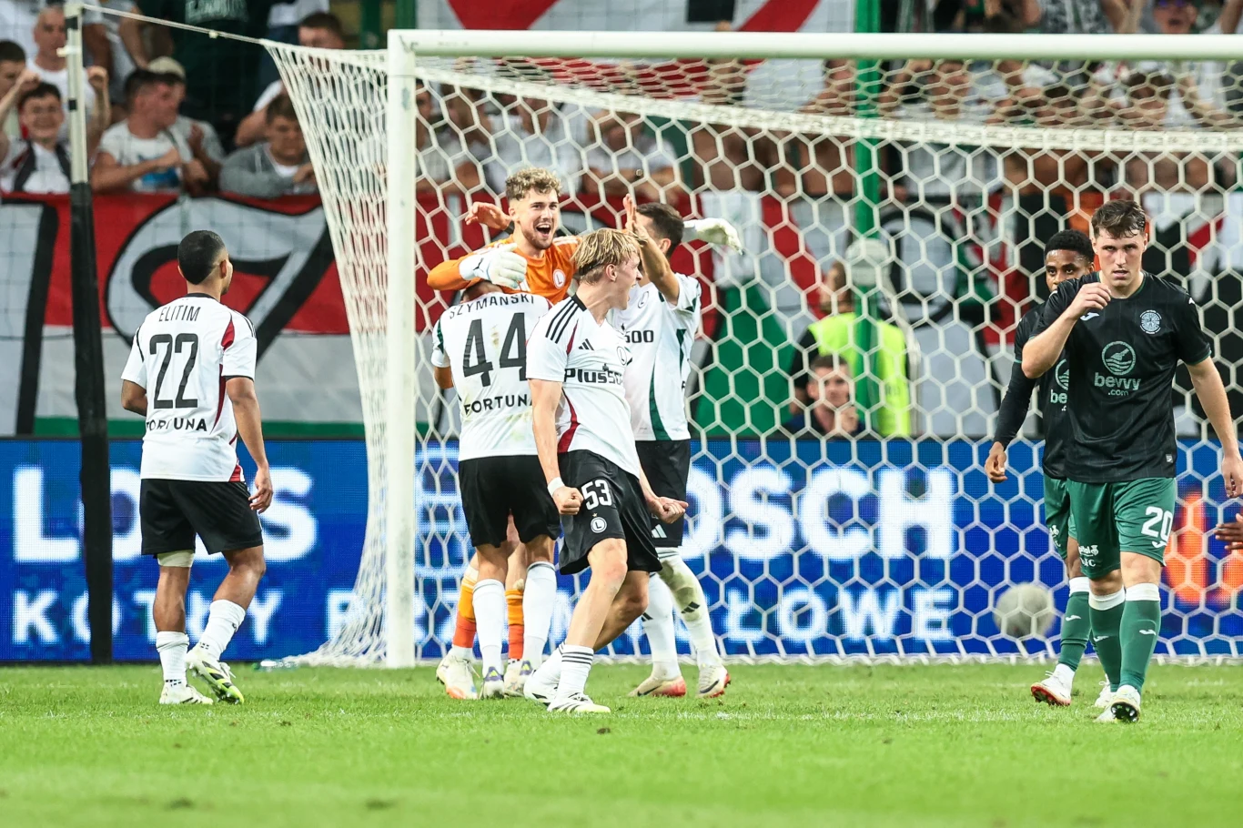 Pikarze Legii Warszawa Piłkarze podczas emocjonującej akcji na murawie stadionu, widoczne wzajemne reakcje i świętowanie oraz grupa kibiców w tle, bramka otoczona siatką, intensywna atmosfera sportowego wydarzenia.