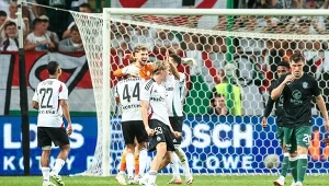 Piłkarze podczas emocjonującej akcji na murawie stadionu, widoczne wzajemne reakcje i świętowanie oraz grupa kibiców w tle, bramka otoczona siatką, intensywna atmosfera sportowego wydarzenia.