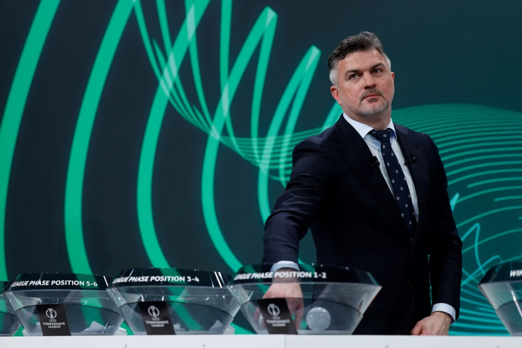 Michał Żewłakow podczas losowania Ligi Konferencji Mężczyzna w garniturze bierze udział w losowaniu, w tle nowoczesna grafika i przezroczyste misy z oznaczeniami pozycji faz grupowych.