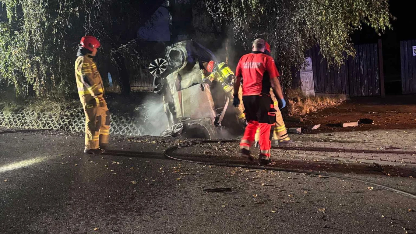 Wypadek śmiertelny w Sierczy Strażacy w specjalnych strojach gaszą wrak samochodu przewróconego na bok na poboczu drogi w nocy, wokół unosi się dym, oświetlenie koncentruje się na miejscu zdarzenia, widoczna intensywna akcja ratownicza.
