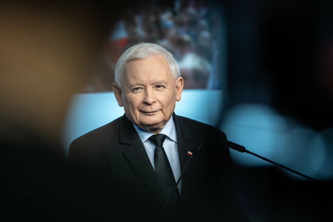 Starszy mężczyzna z siwymi włosami w garniturze i krawacie stoi przy mównicy, tło rozmyte, wskazujące na obecność widowni lub tłumu.