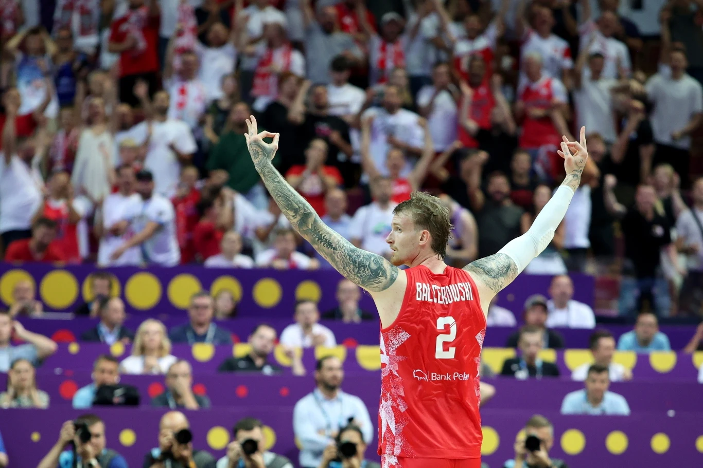 Koszykarz w czerwonym stroju z numerem 2 unosi ręce w geście tryumfu, w tle kibice oraz fotografowie na trybunach, atmosfera entuzjazmu i sportowych emocji.