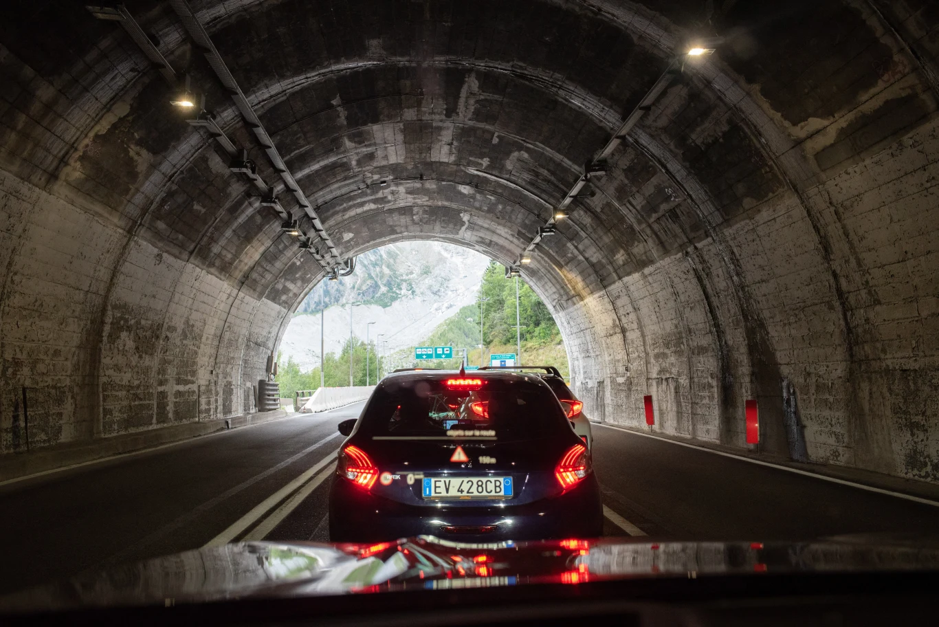 Tunel pod Mont Blanc w Alpach nieczynny do grudnia. Będą utrudnienia Samochody zatrzymane w tunelu, widok z wnętrza samochodu na tył czarnego auta z trójkątem ostrzegawczym oraz światłem dziennym wpadającym przez drugi koniec tunelu, w tle widoczne fragmenty gór i zieleni.