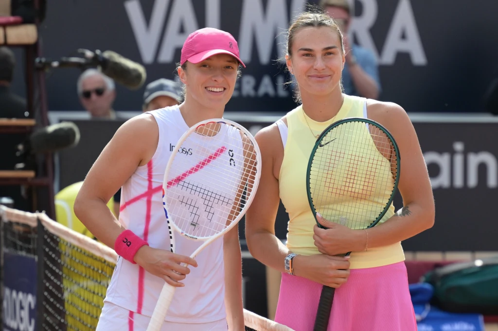 Dwie zawodniczki tenisowe stojące obok siebie przy siatce, obie ubrane w sportowe stroje i trzymające rakiety, w tle widoczne elementy kortu i sprzęt medialny.