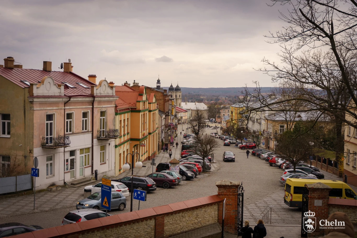Ulica Lubelska przy sanktuarium maryjnym na Górze Chełmskiej. Widok na rynek Panorama miejskiej ulicy w Chełmie z zabytkowymi kamienicami po lewej stronie, zaparkowanymi samochodami oraz widocznym w oddali kościołem.