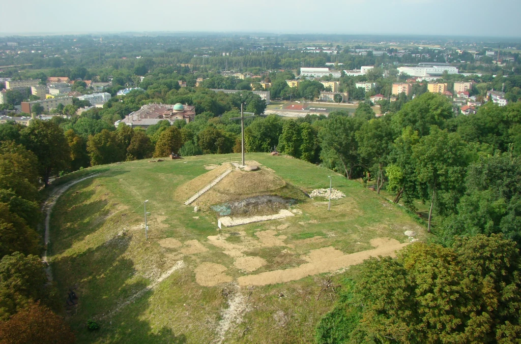 Wzgórze z dużym krzyżem pośrodku, otoczone zielenią drzew, wyraźnie widoczne ścieżki na trawie, w tle panorama miasta z licznymi budynkami.