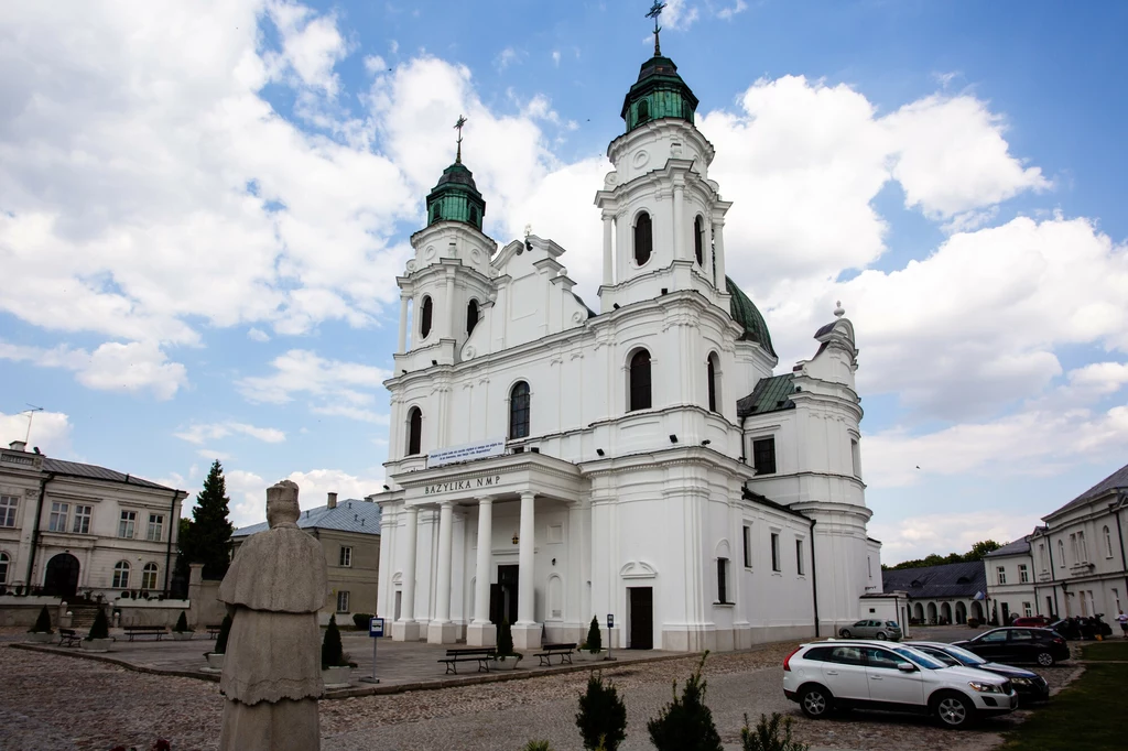 Barokowy kościół z dwoma wieżami i zielonymi kopułami, w tle błękitne niebo z chmurami, przed świątynią kilka zaparkowanych samochodów oraz rzeźba postaci po lewej stronie.