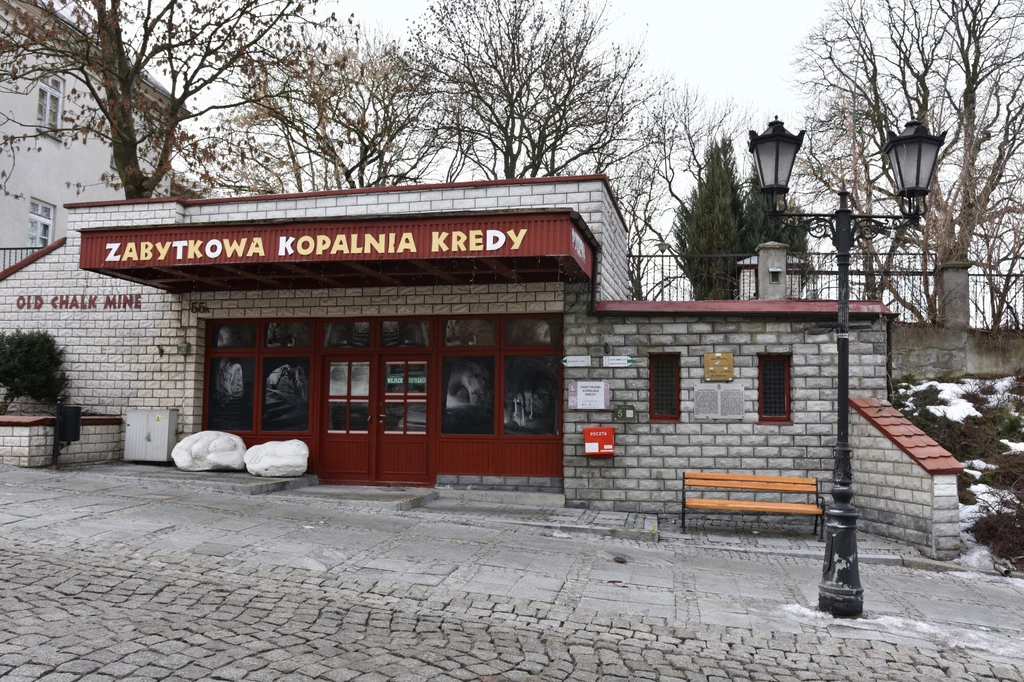 Wejście do zabytkowej kopalni kredy z charakterystycznym czerwonym szyldem, licznymi tablicami informacyjnymi, szerokimi drewnianymi drzwiami i brukowaną nawierzchnią wokół budynku. Przy wejściu stoją dwie białe bryły oraz widoczna jest ławka i stylowa...