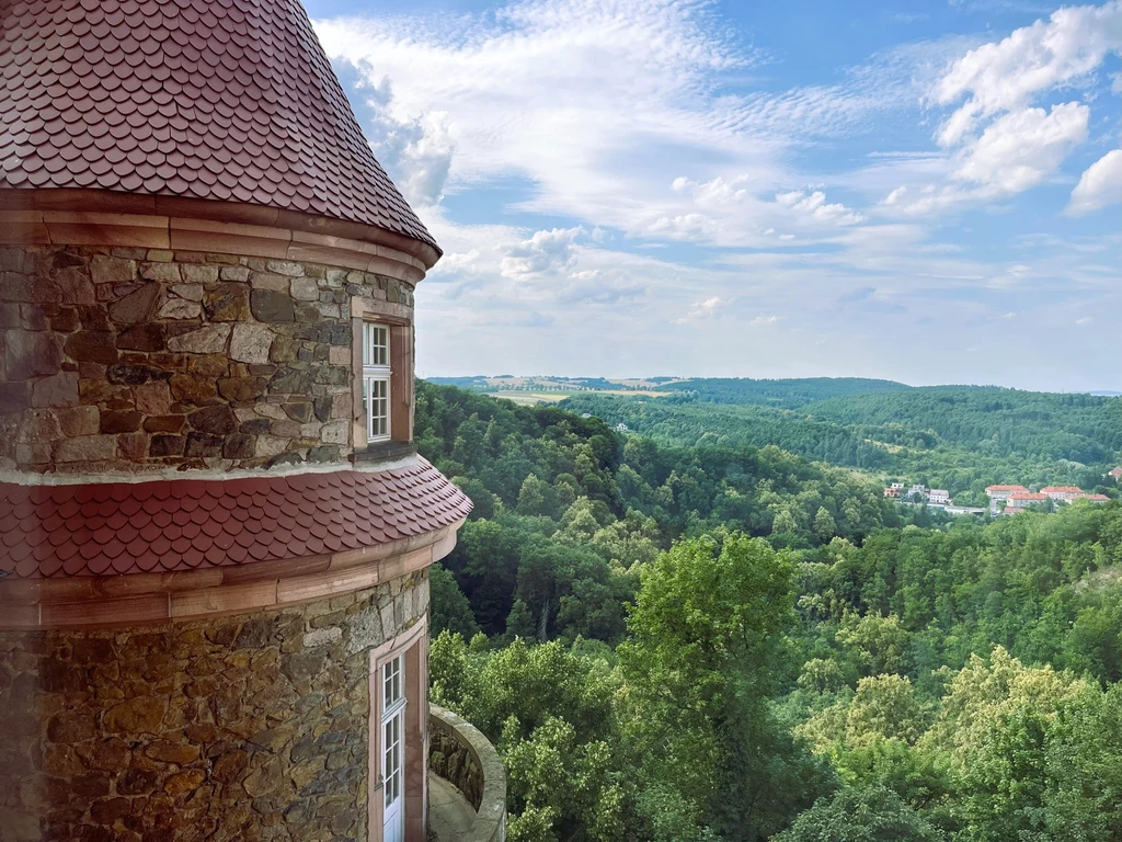 Widoki z zamkowych okien są zachwycające Kamienna baszta zamku z czerwonym dachem i oknami na tle rozległego, zielonego lasu oraz panoramiczny widok na wzgórza i zabudowania oddalone w pejzażu.