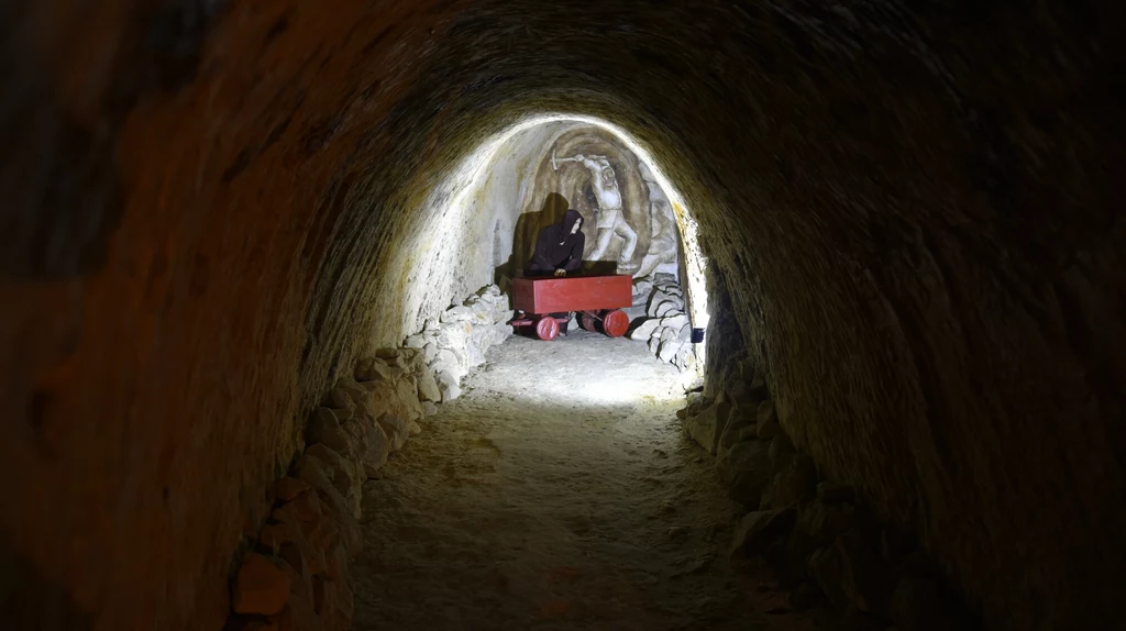 Kamienny tunel oświetlony w centrum, w głębi widoczny czerwony wózek górniczy oraz figura stojącej postaci przy żłobionej ścianie.