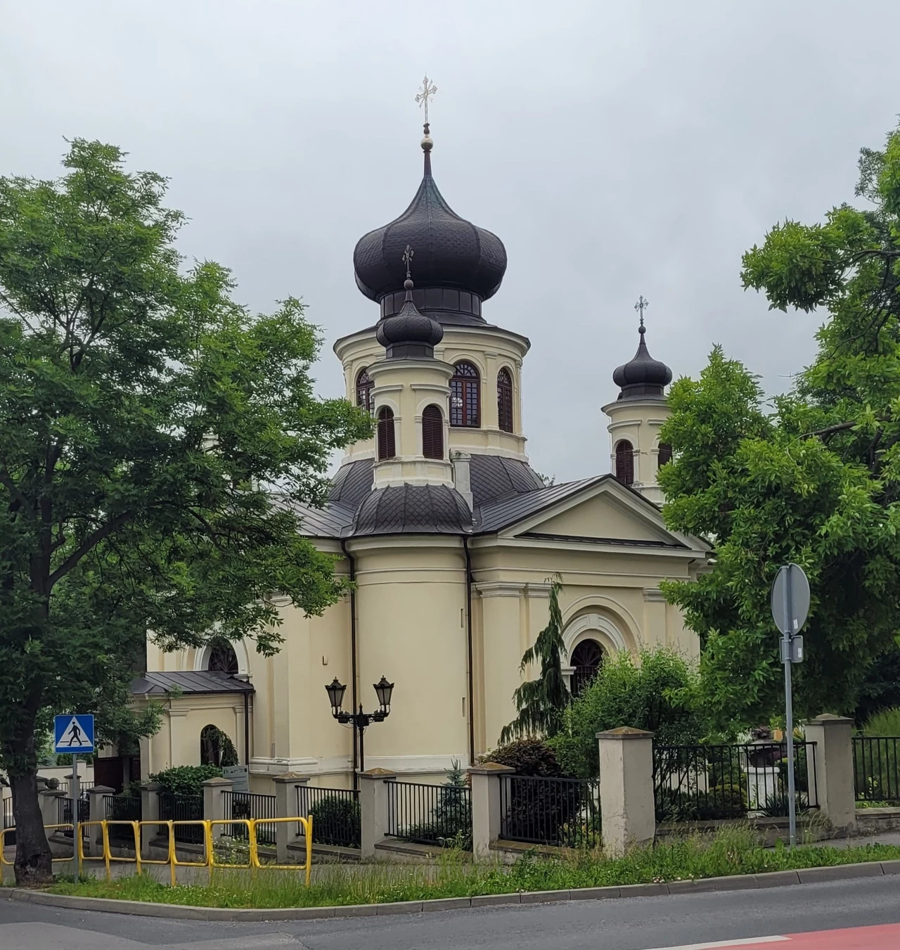 Cerkiew prawosławna o charakterystycznej architekturze z dużą cebulastą kopułą, otoczona drzewami, z widocznym ogrodzeniem i elementami infrastruktury miejskiej, położona przy ulicy
