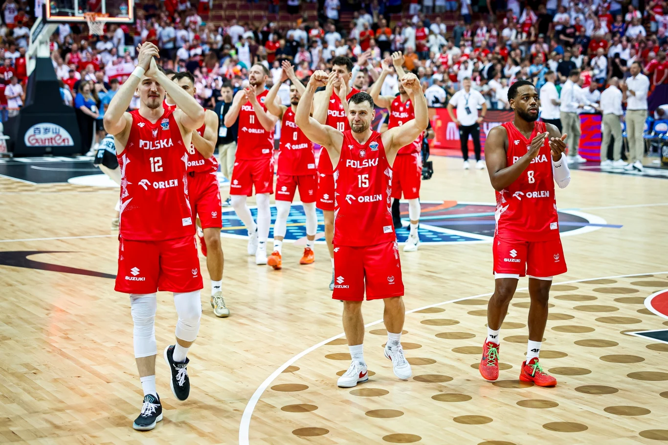 Polacy wykonali świetną robote i pokonali Słowenię Polscy koszykarze ubrani w czerwone stroje drużynowe z napisem POLSKA na tle hali sportowej, uniesione ręce w geście podziękowania kibicom, na parkiecie widoczne logo i sędziowie oraz publiczność na trybunach.