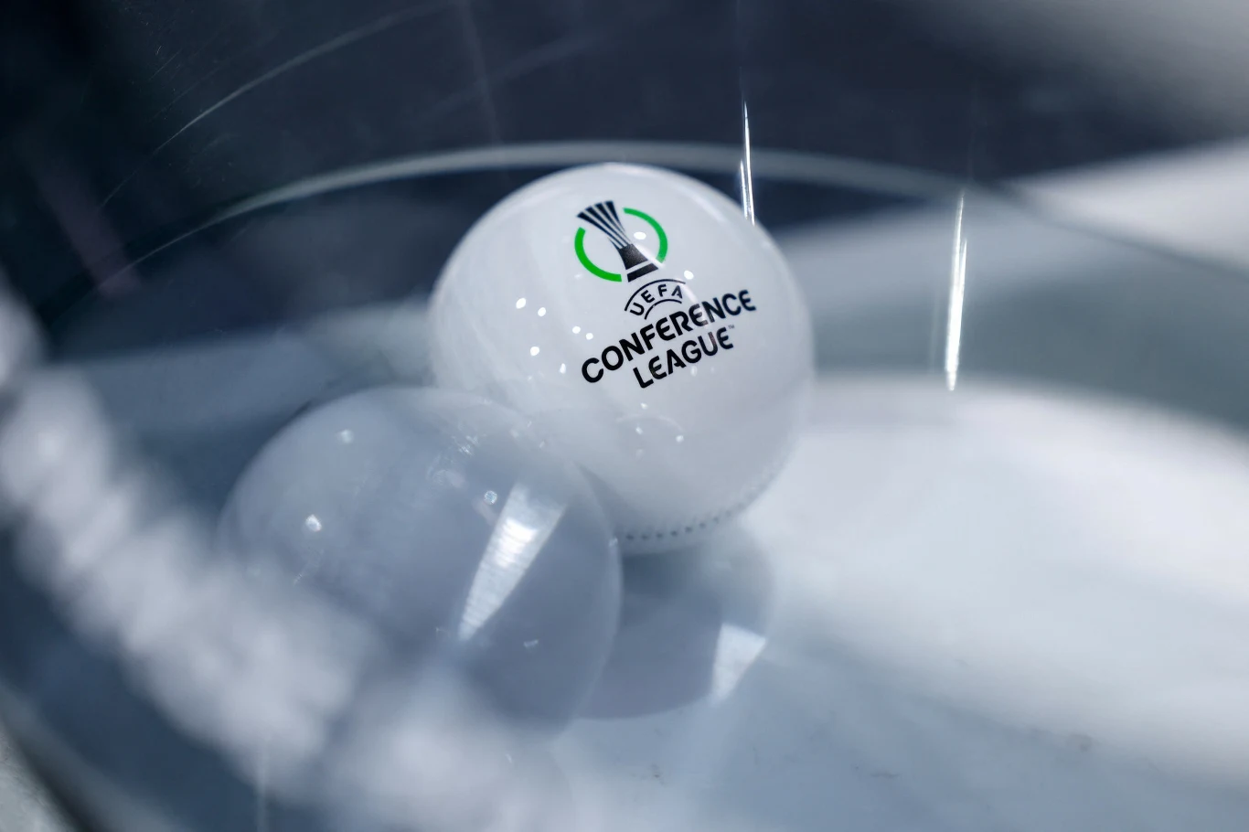 Biała kula z logo UEFA Conference League umieszczona w przezroczystym naczyniu, obok znajduje się druga, przezroczysta kula. Główna kula jest wyraźnie oznakowana i wskazuje na związek z rozgrywkami piłkarskimi.