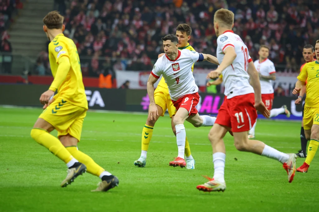 Rober Lewandowski i Karol Świderski w trakcie meczu Polska - Litwa Zawodnicy dwóch reprezentacji w trakcie meczu piłki nożnej, w centrum piłkarz w białej koszulce i czerwonych spodenkach walczący o piłkę z przeciwnikiem w żółtym stroju, w tle kibice na trybunach.