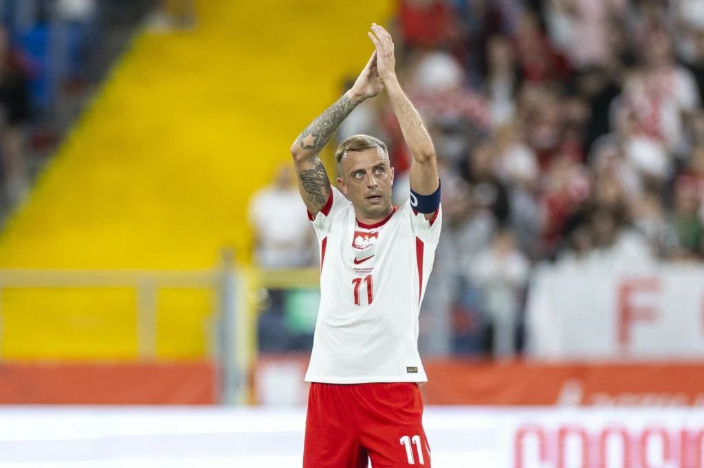 Piłkarz w białoczerwonym stroju z numerem 11 podnosi ręce do góry, klaszcząc w stronę publiczności na stadionie; w tle rozmyta widownia i fragment boiska.