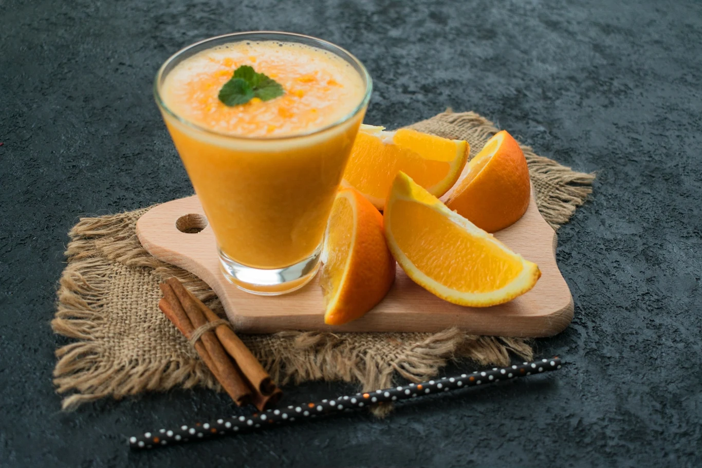 Sok, a w zasadzie smoothie przygotowane samemu w domu będzie najzdrowszym wyborem Szklanka świeżego soku pomarańczowego ozdobiona listkiem mięty, otoczona kilkoma kawałkami pomarańczy na drewnianej desce, obok znajdują się dwa laski cynamonu i rurki do picia.