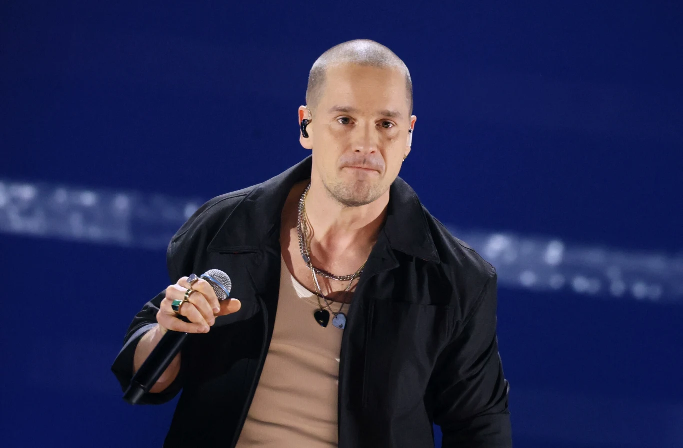 Krzysztof Zalewski pokazał swojego ojca na Instagramie Mężczyzna o ogolonej głowie w jasnej koszulce i czarnej kurtce, trzyma mikrofon na scenie. W tle jednolite, granatowe tło z widocznym, jasnym pasem świetlnym. Postać nosi łańcuszki na szyi oraz pierścionki na palcach.