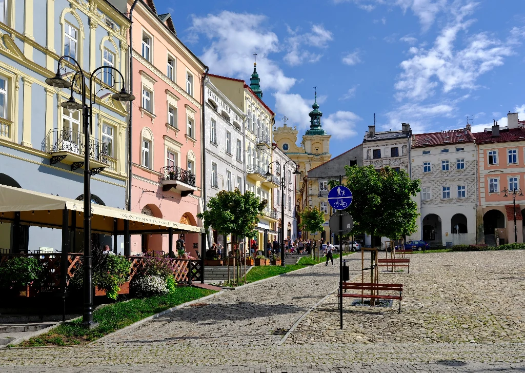 Na przemyskim Rynku znajdują się przede wszystkim kamienice miejskie z podcieniami Kolorowe kamienice z podcieniami i balkonami ciągną się wzdłuż brukowanej ulicy, po której rozstawione są ławki i latarnie, a w tle widoczne są zielone wieże kościoła, na placu rosną drzewa i widoczni są przechodnie.