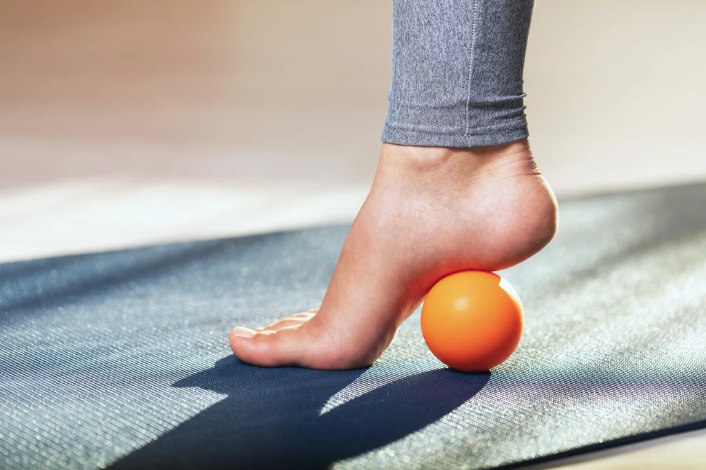 Regularne rolowanie stóp przynosi wiele korzyści Stopa osoby w szarej legginsie masująca się na pomarańczowej piłeczce do masażu, leżącej na macie treningowej.