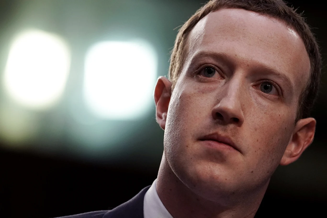 Mark Zuckerberg Mark Zuckerberg w garniturze patrzy w bok, lekko oświetlona twarz, ciemne tło z dwoma jasnymi światłami.