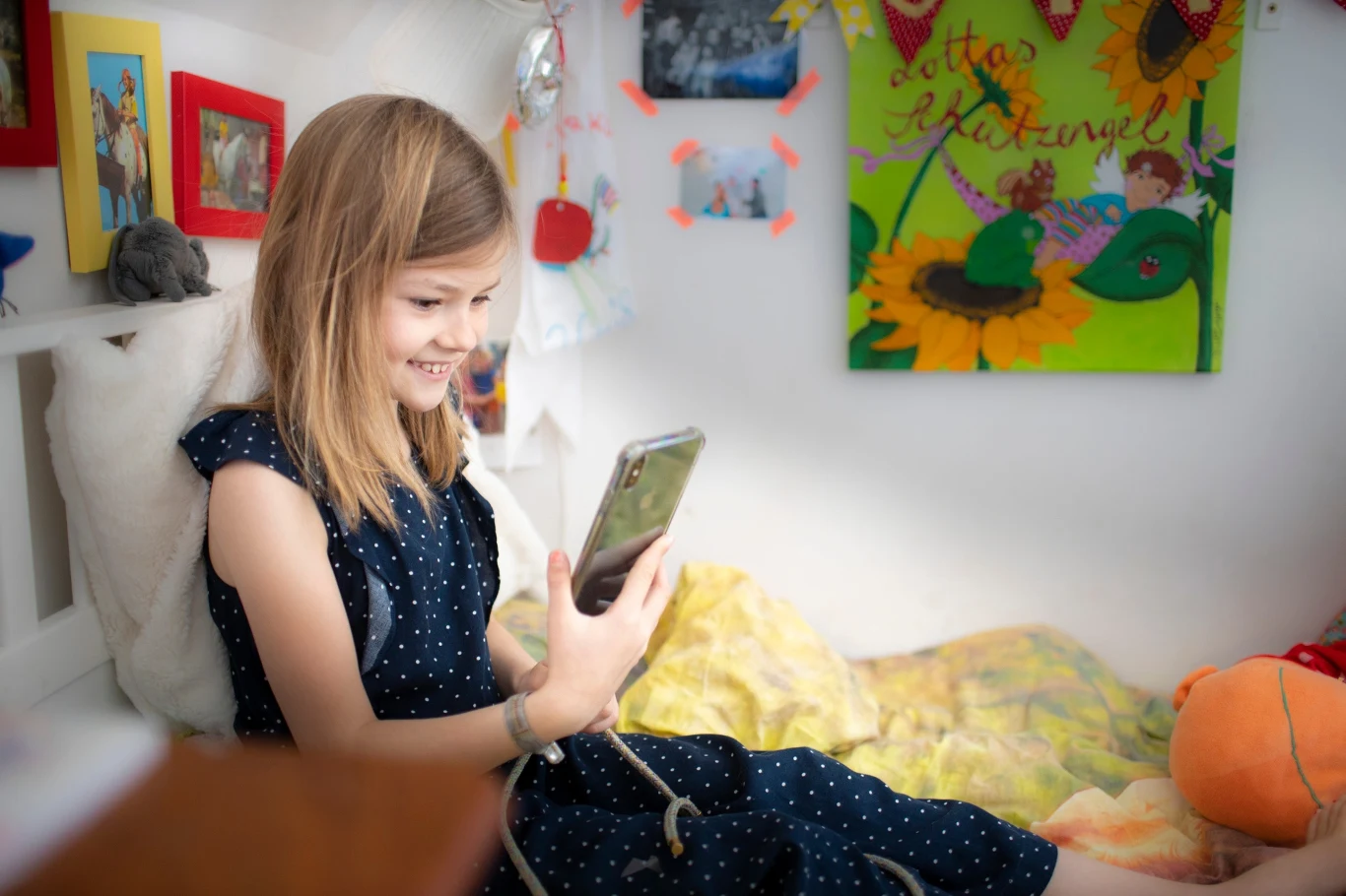 To proszenie się o problemy. Dzieci coraz rzadziej wychodzą z domu na świeże powietrze Dziewczynka w granatowej sukience z uśmiechem patrzy na smartfon, siedząc na łóżku w kolorowo udekorowanym pokoju dziecięcym, za nią widoczne są rysunki oraz pluszowa zabawka.