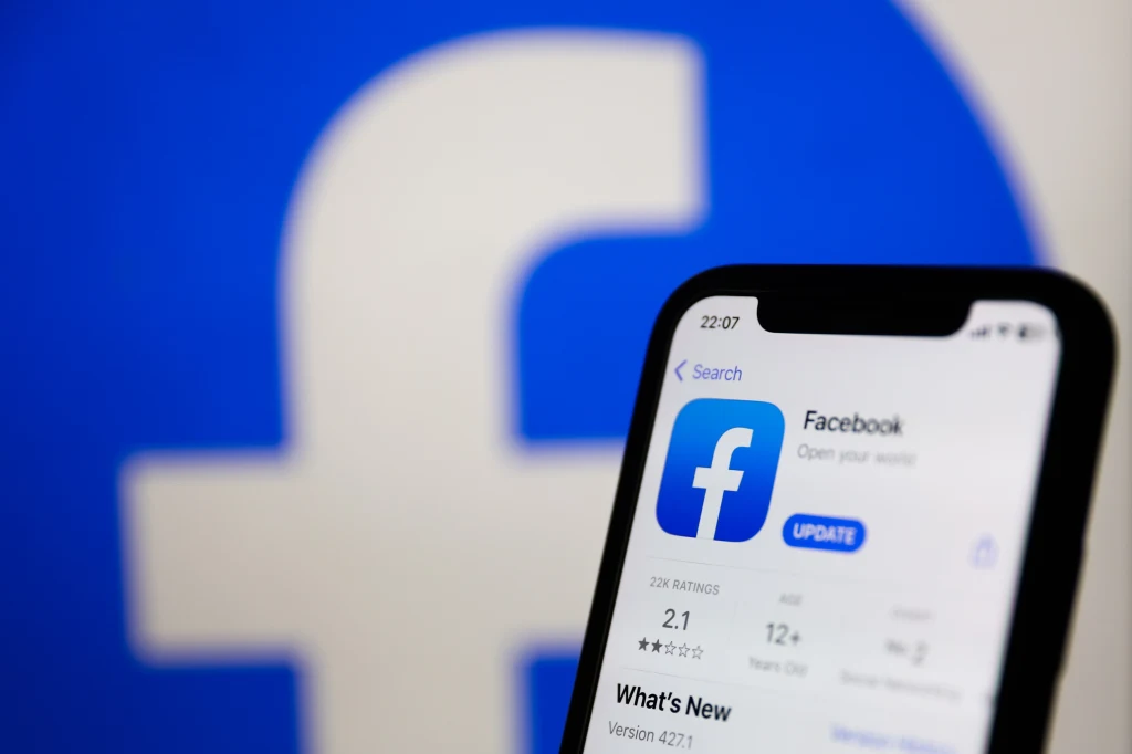Facebook rządzi pod kątem liczby aktywnych użytkowników na świecie. Ekran smartfona z otwartą stroną aktualizacji aplikacji Facebook na tle dużego, rozmytego logo Facebooka w kolorze niebieskim.