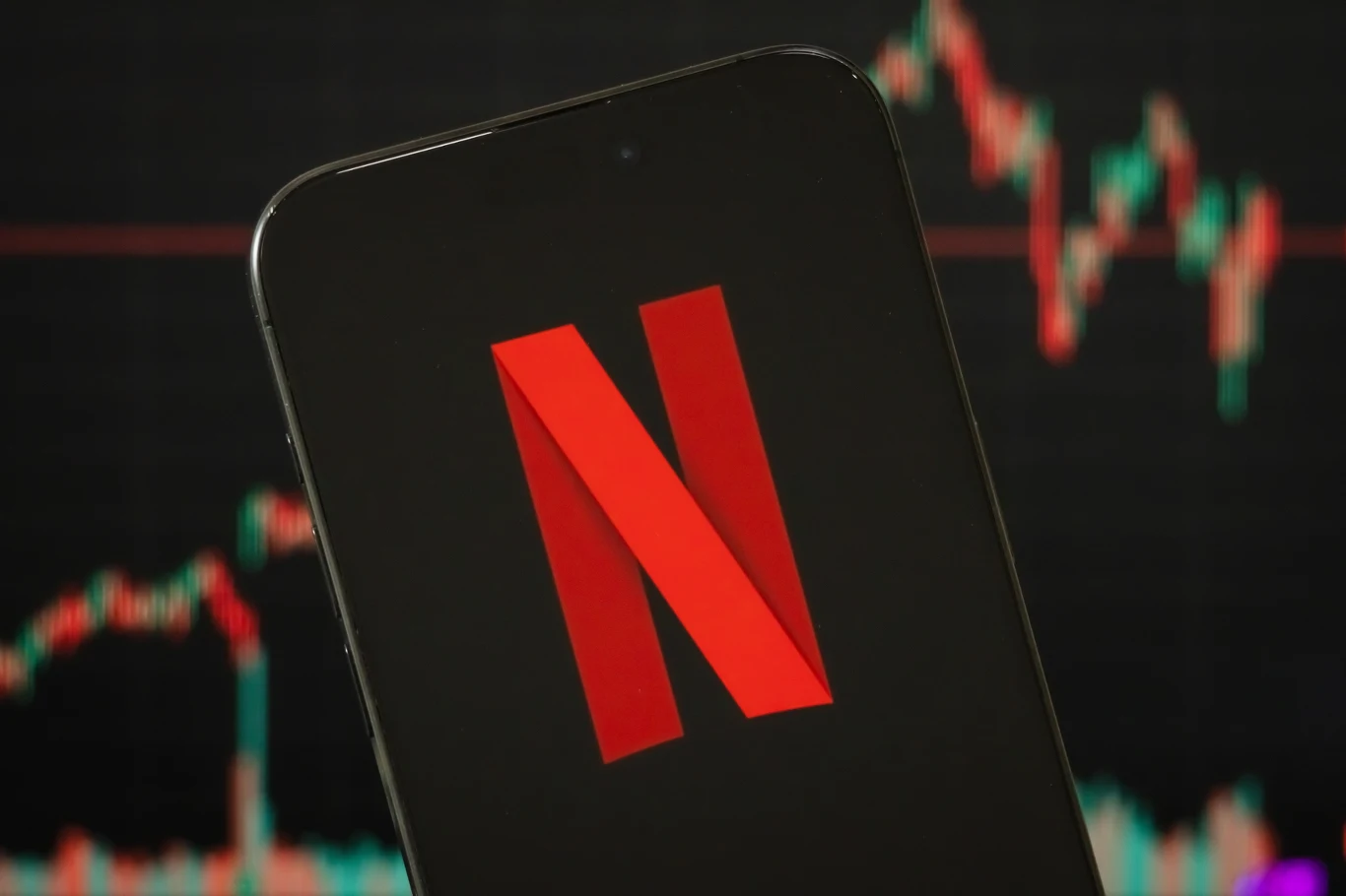 Netflix Logo Netflix wyświetlone na ekranie smartfona, w tle rozmazany wykres giełdowy obrazujący zmiany wartości akcji.