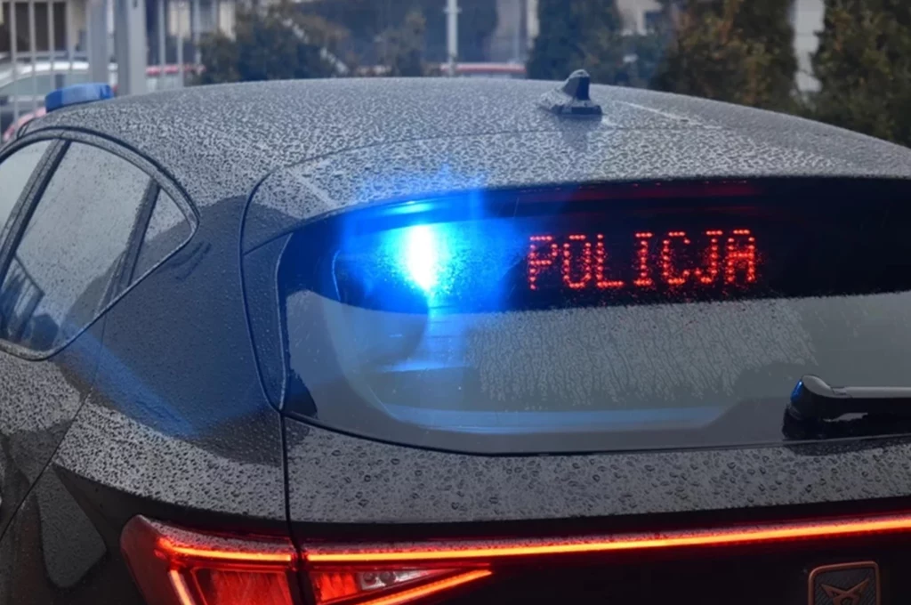Radiowóz policyjny z włączonym niebieskim światłem sygnalizacyjnym, na tylnej szybie widoczny jest cyfrowy napis 'POLICJA', samochód mokry od deszczu, w tle nieostre elementy miejskiego otoczenia.