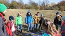 Grupa dzieci i dorosły stoją w kręgu na trawie, uczestnicząc w grze edukacyjnej z planszami rozłożonymi na ziemi, wokół jesienne drzewa i otwarta przestrzeń parku.