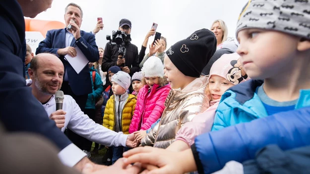 Grupa dzieci w kolorowych kurtkach i czapkach stoi w rzędzie, ściskając dłonie z dorosłym mężczyzną, wokół obecni są inni dorośli, część osób dokumentuje wydarzenie aparatami i telefonami.