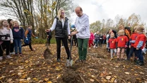 Grupa dzieci i dorosłych uczestniczy w sadzeniu drzew na jesiennym, liściastym terenie, dwie osoby na pierwszym planie wspólnie trzymają łopatę i sadzonkę, wokół zgromadzona grupa obserwujących.