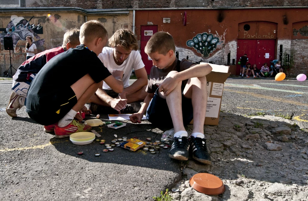 Podwórkowe gangi nigdy się nie nudziły Grupa dzieci i młodych osób siedzi na asfalcie, pochylając się nad planszą do gry i rozrzuconymi wokół elementami. W tle stara ceglana ściana z kolorowym graffiti i innymi dziećmi, które również spędzają czas na podwórku.