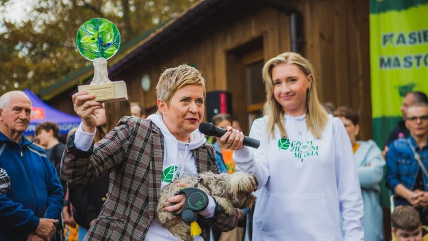 Kobieta w marynarce i białej bluzie trzymająca statuetkę z zielonym elementem oraz psa, obok niej druga kobieta z mikrofonem, w tle zgromadzeni ludzie oraz banery wydarzenia.
