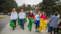Grupa młodych ludzi i dorosłych idzie chodnikiem w otoczeniu drzew, trzymając worki przeznaczone do segregacji odpadów; w tle widoczna zabudowa oraz inne osoby uczestniczące w wydarzeniu.