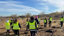 Grupa osób w odblaskowych kamizelkach idzie po polu, na którym przygotowano już dołki i rozsypano ziemię, niebo jest częściowo zachmurzone, w tle widać drzewa i krzewy, na ziemi leży łopata.
