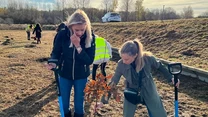 Dwie kobiety sadzą młode drzewko na otwartym terenie, obok nich leżą łopaty, w tle widoczne inne osoby uczestniczące w akcji sadzenia oraz samochód zaparkowany na wzniesieniu.
