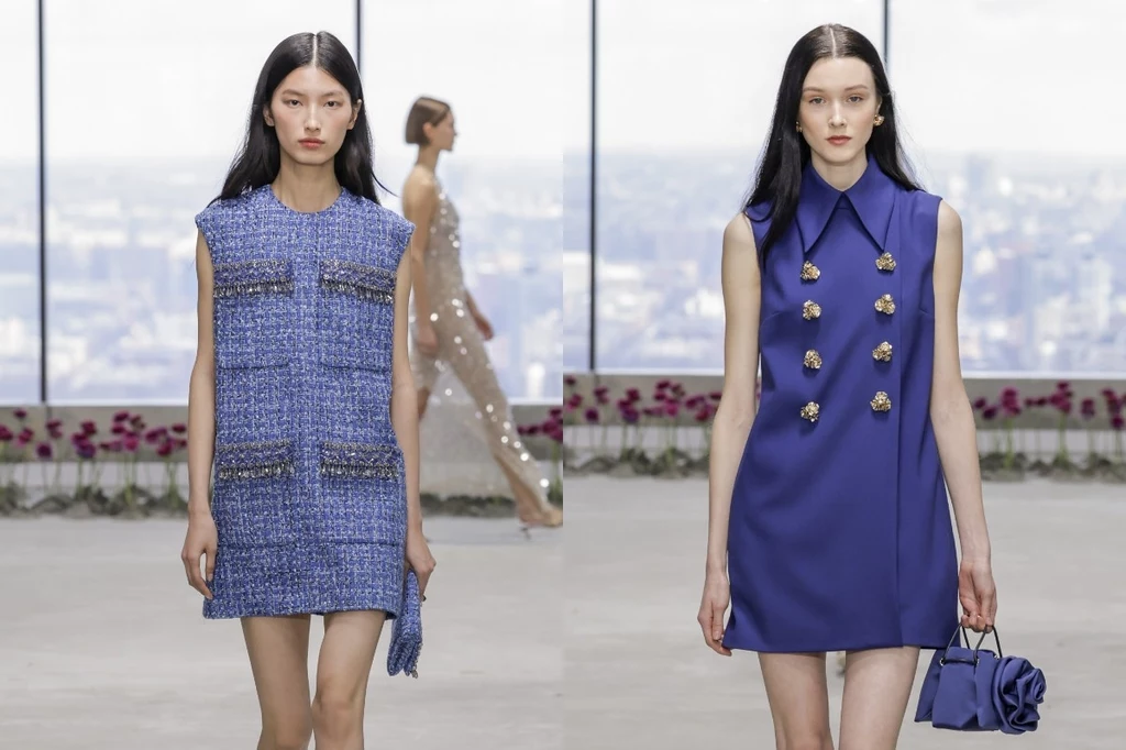Carolina Herrera autumn/winter 2025-2026 Dwie młode kobiety prezentują nowoczesne sukienki w odcieniach niebieskiego na tle dużych okien z panoramą miasta, jedna trzyma torebkę; w tle widoczna jest sylwetka innej modelki.