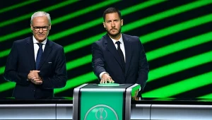 Dwóch mężczyzn ubranych w eleganckie garnitury stoi przy podium z logo UEFA Conference League, jeden z nich naciska przycisk, tło w zielone ukośne linie.