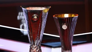 Dwa puchary sportowe ustawione obok siebie, jeden z fakturowanym metalicznym wykończeniem i okrągłym emblematem, drugi smuklejszy z gładkimi pionowymi żłobieniami i złotym wnętrzem.