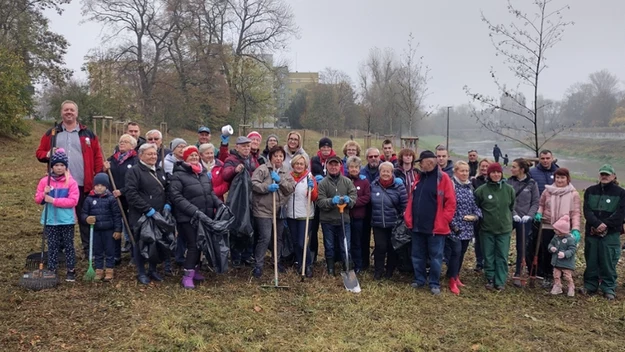 Grupa ludzi różnych pokoleń, ubranych w kurtki i ciepłe ubrania, zgromadzona na trawniku podczas wspólnej akcji sprzątania. Część osób trzyma worki na śmieci, łopaty i inne narzędzia do porządkowania terenu. W tle drzewa, rzeka i jesienna aura.