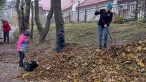 Grupa osób sprząta liście na skarpie w parku lub przy ulicy, dzieci i dorośli zbierają liście do worków, używając grabi, otoczenie jesienne z gołymi drzewami i budynkiem w tle.